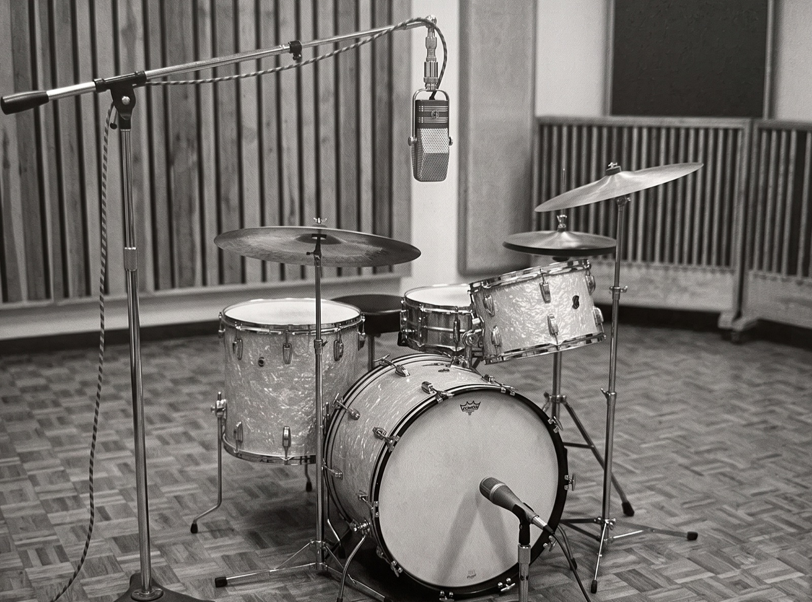 Black and white photograph of a vintage four-piece drum kit with a pearl wrap finish, recorded with only two microphones: a single large ribbon microphone on a tall boom stand above the kit, and one dynamic microphone low in front of the kick drum, in a wood-panelled studio with a parquet floor.