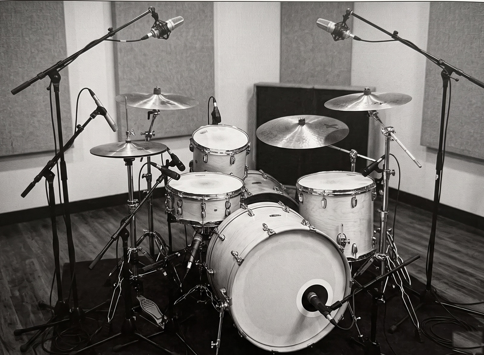 Black and white photograph of a modern studio drum kit rigged for dense close-miking: a dynamic microphone at the kick drum port, clip-on microphones at the snare and toms, a pencil condenser at the hi-hat, and a stereo pair of condenser overheads on tall boom stands, in a treated live room with acoustic panels.